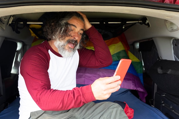 Schwuler Mann mittleren Alters sitzt in einem Campingbus mit LGBTI-Flagge und gestikuliert mit einem Smartphone