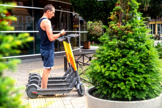 Scooter eléctrico y teléfono móvil. Hombre que usa el servicio de alquiler de vehículos e con teléfono inteligente en la calle y el parque urbanos de la ciudad. Joven alquila un transporte ecológico en verano. estacionamiento para patineta eléctrica.