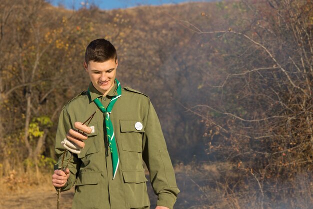 Scout preparándose para cocinar salchichas en un fuego de pie mirando hacia el humo mientras sostiene las salchichas enhebradas en una brocheta o palo