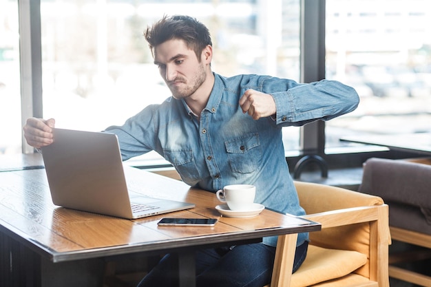 Sde view retrato de agressivo jovem empresário infeliz em camisa jeans azul está sentado no café e de mau humor estão prontos para socar um trabalhador através de uma webcam com o punho levantado com cara de bravo.