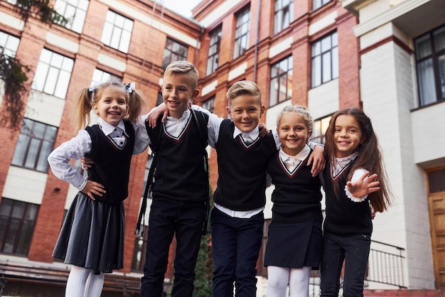 Se divertindo e se abraçando. grupo de crianças com uniforme escolar que fica junto ao ar livre, perto do prédio da educação.