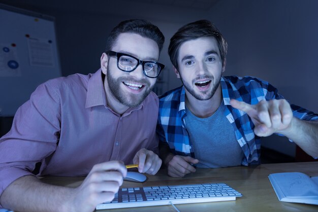Se divertindo. Homens bonitos alegres e positivos sentados à mesa e rindo enquanto usam um computador
