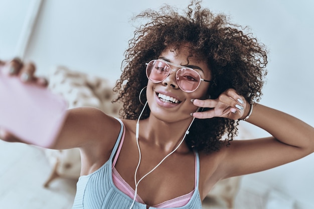 Se divertindo. Mulher jovem e atraente africana ouvindo música e sorrindo enquanto tira uma selfie em casa