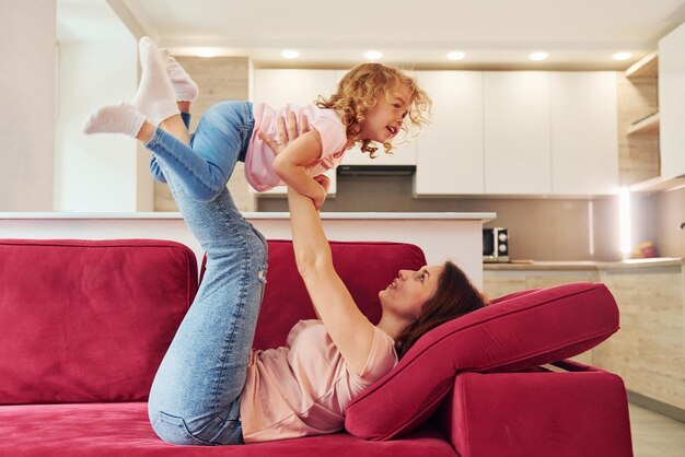Se divertindo no sofá vermelho Jovem mãe com sua filha em roupas casuais juntos dentro de casa