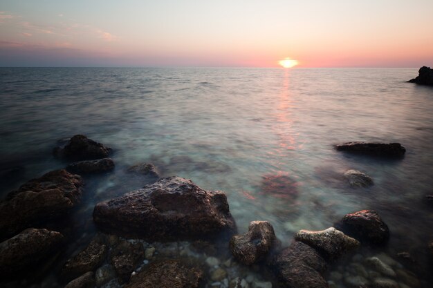 Seascape da costa de águas do mar ainda, litoral rochoso e pôr do sol rosa romântico