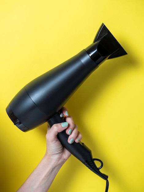 Secador de cabelo preto na mão de uma mulher em um fundo amarelo. Produto para cuidados com o cabelo