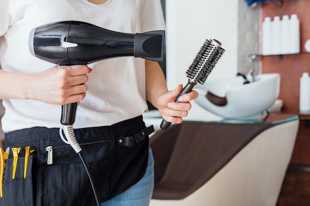 Foto secador de pelo, cepillo de peine en manos de peluquería contra salón de belleza, interior de peluquería, lavabo. herramientas profesionales de peluquería.