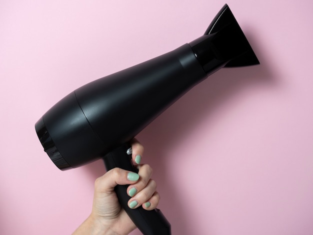 Secador de pelo negro en la mano de una mujer sobre un fondo rosa. Producto para el cuidado del cabello.