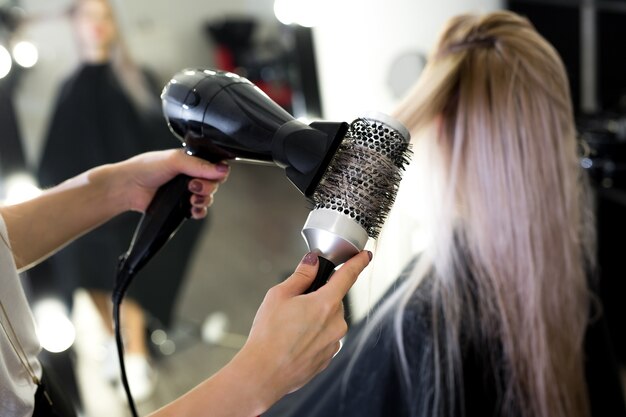 Secagem de cabelo no estúdio de cabeleireiro. estilista de cabeleireira  seca o cabelo com secador de cabelo e escova redonda ruiva de uma mulher em  um salão de beleza.