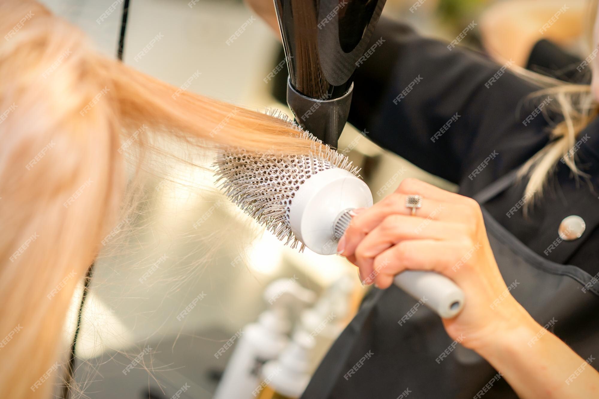 Secagem de cabelo no estúdio de cabeleireiro. estilista de cabeleireira  seca o cabelo com secador de cabelo e escova redonda ruiva de uma mulher em  um salão de beleza.