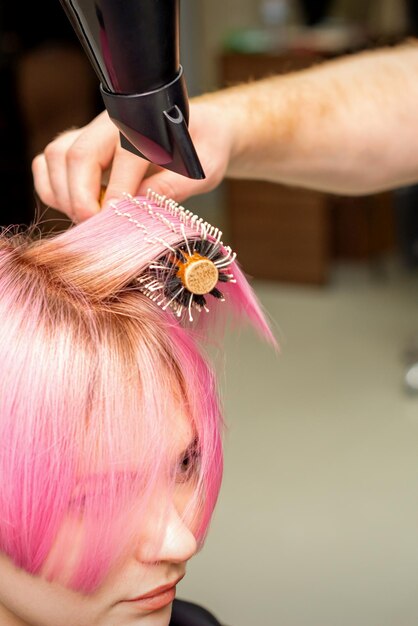 Secagem de cabelo rosa curto de jovem mulher caucasiana com um secador de cabelo preto e escova redonda preta pelas mãos de um cabeleireiro masculino em um salão de cabeleireiro close-up