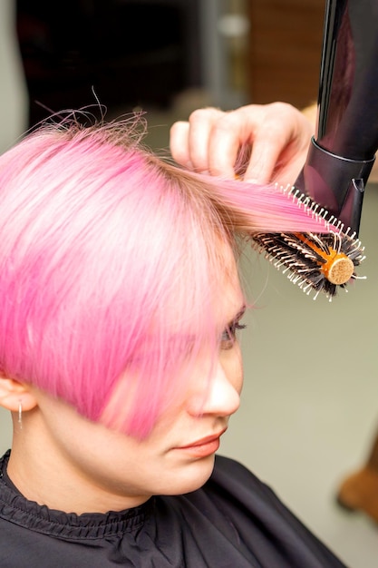 Secagem de cabelo rosa curto de jovem mulher caucasiana com um secador de cabelo preto e escova redonda preta pelas mãos de um cabeleireiro masculino em um salão de cabeleireiro close-up