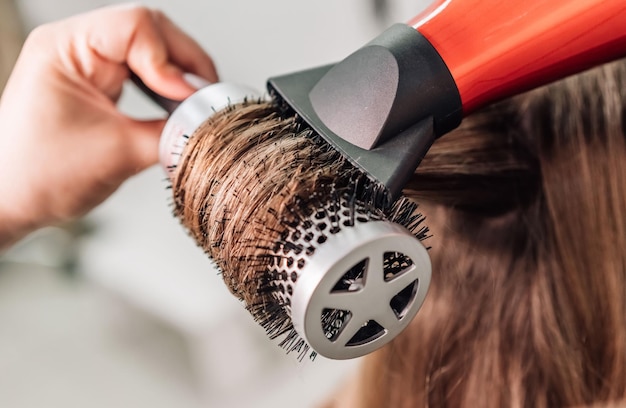 Secagem de cabelo no estúdio de cabeleireiro. estilista de cabeleireira  seca o cabelo com secador de cabelo e escova redonda ruiva de uma mulher em  um salão de beleza.