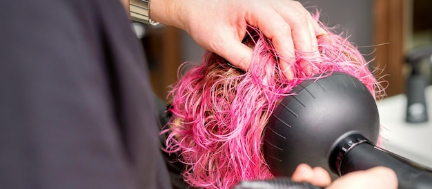 Secando o penteado curto rosa bob de uma jovem mulher caucasiana com um secador de cabelo preto com a escova pelas mãos de um cabeleireiro masculino em um salão de cabeleireiro close-up