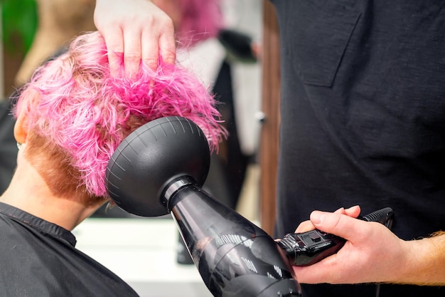 Secando o penteado curto rosa bob de uma jovem mulher caucasiana com um secador de cabelo preto com a escova pelas mãos de um cabeleireiro masculino em um salão de cabeleireiro close-up