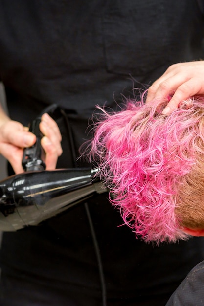 Secando o penteado curto rosa bob de uma jovem mulher caucasiana com um secador de cabelo preto com a escova pelas mãos de um cabeleireiro masculino em um salão de cabeleireiro close-up