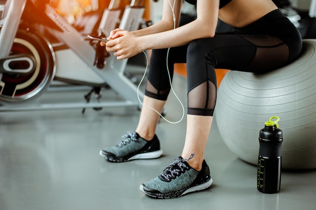 Seção baixa de mulher ouvindo música no telefone móvel enquanto está sentada sobre uma bola de fitness no ginásio