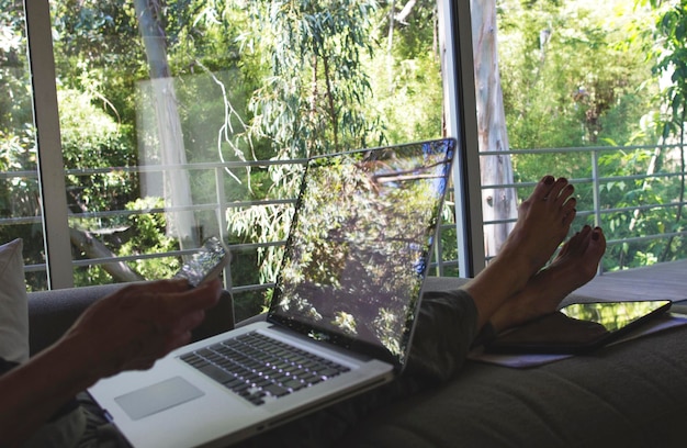 Foto seção baixa de mulher usando telefone móvel enquanto está sentada com laptop em casa