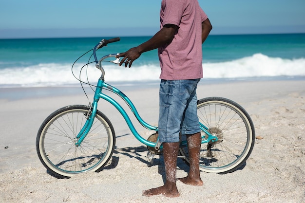 Seção baixa de um homem afro-americano sênior de pé descalço na praia com céu azul e mar ao fundo, segurando uma bicicleta azul