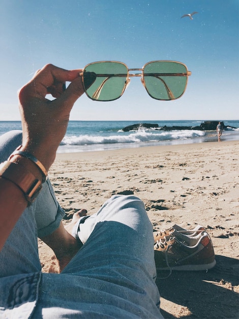 Foto seção baixa de um homem segurando óculos de sol enquanto relaxa na praia contra o céu