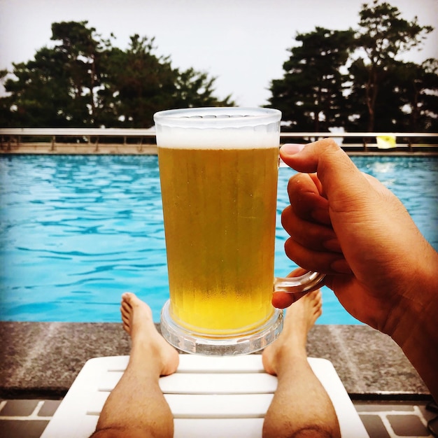 Seção baixa de um homem segurando um copo de cerveja enquanto está sentado à beira da piscina