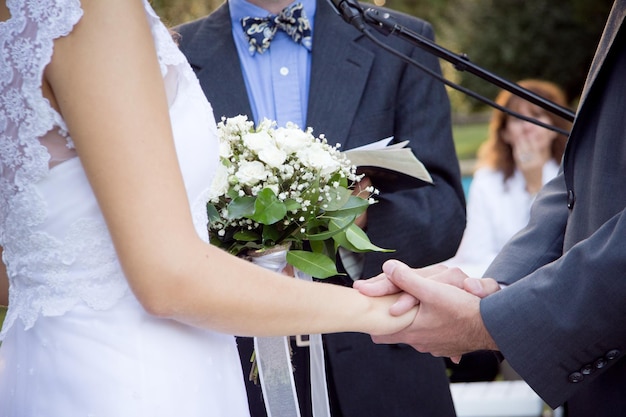 Seção do meio da noiva e do noivo de mãos dadas pelo padre durante a cerimônia de casamento