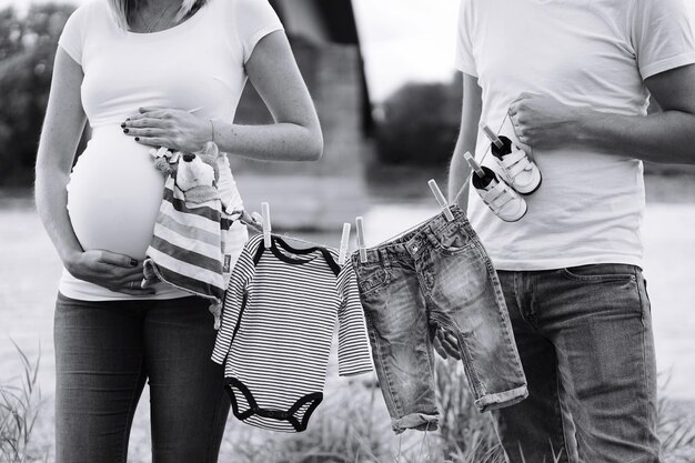 Foto seção média de mulher grávida e homem segurando roupas de bebê no campo