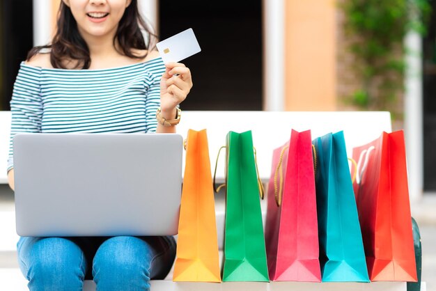 Foto seção média de mulher segurando cartão de crédito enquanto usa laptop por sacos de compras coloridos na cidade