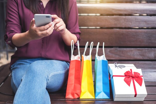 Foto seção média de mulher usando telefone inteligente enquanto está sentada com sacos de compras coloridos em um banco