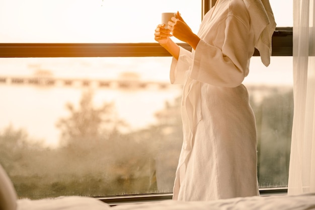 Seção média de uma mulher segurando uma xícara de café pela janela de um quarto de hotel