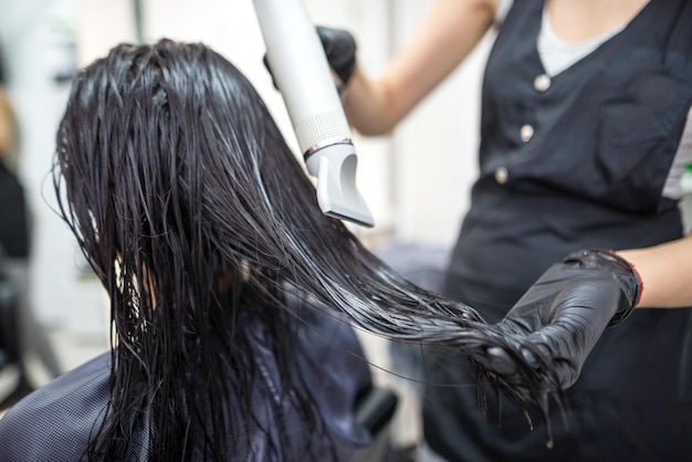 Foto secar el cabello largo y oscuro con un secador de pelo, un peluquero profesional seca el cabello de un cliente en el salón de belleza, peinado, cuidado del cabello, servicio de moda.