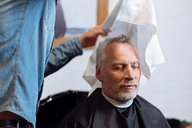 Secar con un paño. Retrato de anciano satisfecho durante la limpieza de su cabello en la peluquería con una toalla especial.