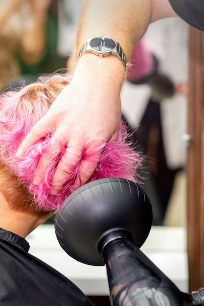 Secar el peinado bob rosa corto de una joven caucásica con un secador de pelo negro con el cepillo a manos de un peluquero masculino en una peluquería de cerca