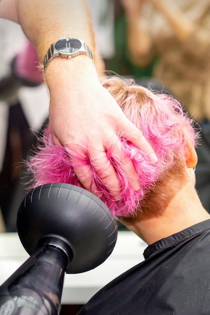 Secar el peinado bob rosa corto de una joven caucásica con un secador de pelo negro con el cepillo a manos de un peluquero masculino en una peluquería de cerca