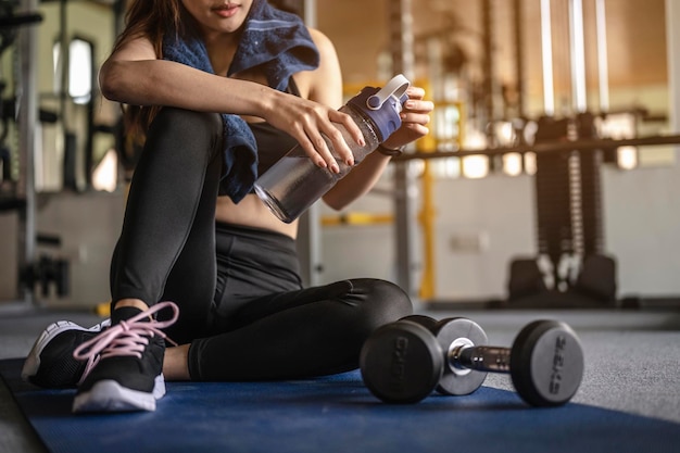 Foto sección baja de una mujer con una botella de agua sentada en una alfombra de ejercicio en el gimnasio