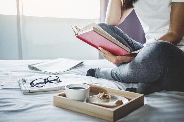 Foto sección baja de una mujer leyendo un libro con comida y bebida en la cama en casa