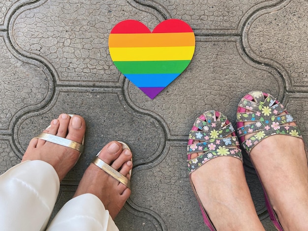 Foto sección baja de una pareja de lesbianas de pie junto a la bandera del arco iris en forma de corazón