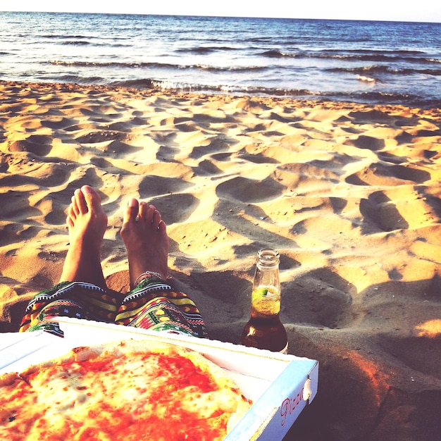 Sección baja de una persona sentada en una playa de arena con pizza y una botella de cerveza