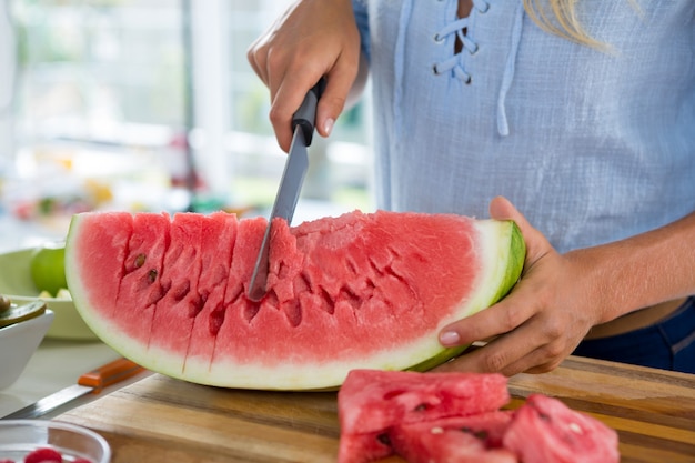 Sección intermedia de mujer cortando frutas sobre una tabla de cortar contra el fondo blanco.