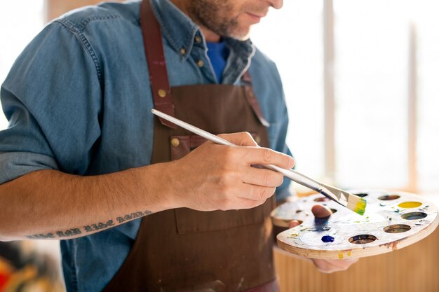 Sección intermedia del pintor contemporáneo en delantal sosteniendo paleta con colores mezclados y pincel durante el trabajo