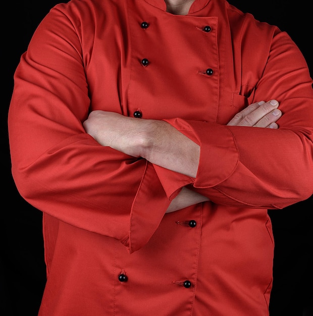 Foto sección media del chef con uniforme rojo de pie con los brazos cruzados contra un fondo negro