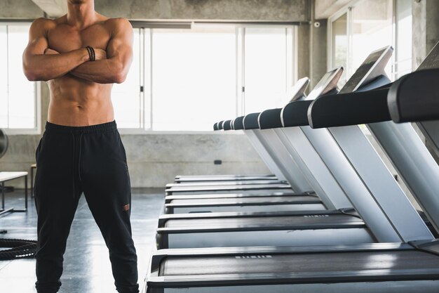 Sección media de un hombre sin camisa de pie junto a las cintas de correr en el gimnasio