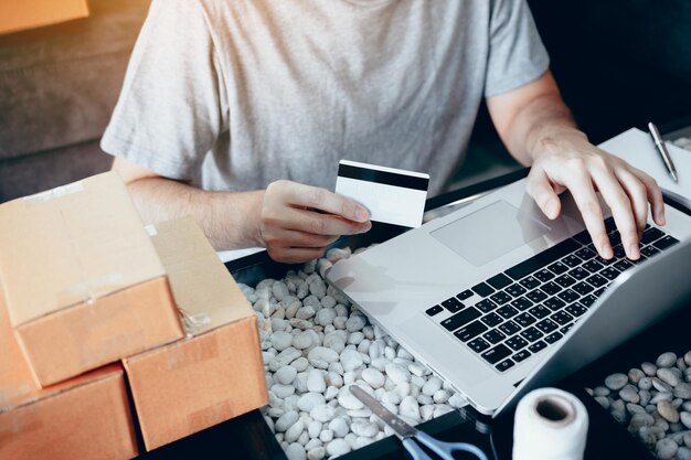 Sección media de un hombre con una tarjeta de crédito mientras usa una computadora portátil con cajas de cartón en la mesa