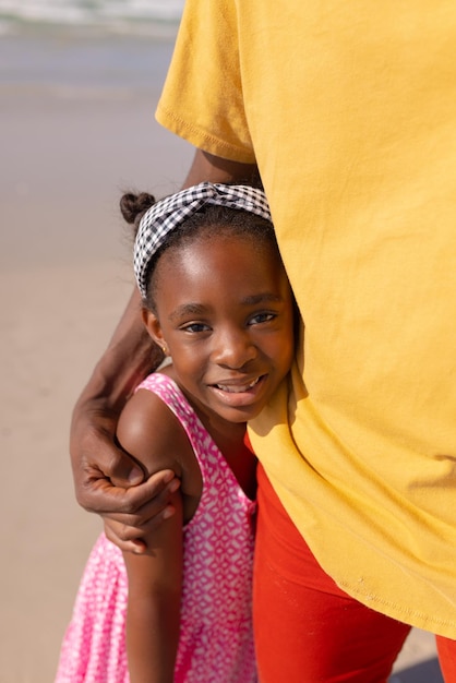 Sección media de un joven afroamericano abrazando a una hija sonriente mientras está de pie en la playa