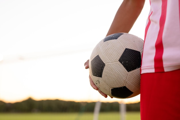 Sección media de un jugador de fútbol masculino caucásico con camiseta roja sosteniendo una pelota de pie contra el cielo claro