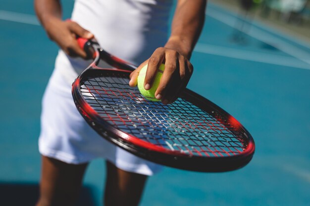 Sección media de una jugadora afroamericana sosteniendo la pelota en una raqueta en una cancha de tenis en un día soleado