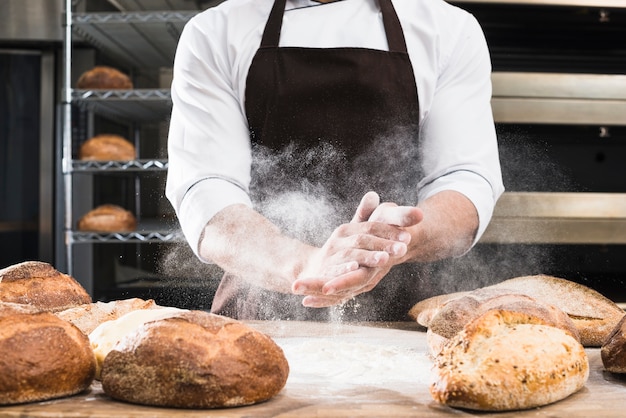 Foto sección media de la mano de un panadero masculino que quita la harina del escritorio de madera con pan horneado