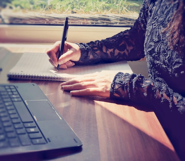 Foto sección media de una mujer escribiendo en un libro con una computadora portátil en la mesa