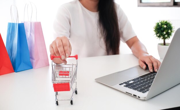 Sección media de una mujer sosteniendo un carrito de compras en miniatura mientras usa una computadora portátil en la mesa