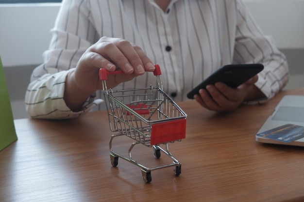 Sección media de una mujer sosteniendo un pequeño carrito de compras mientras está sentada con el teléfono en la mesa